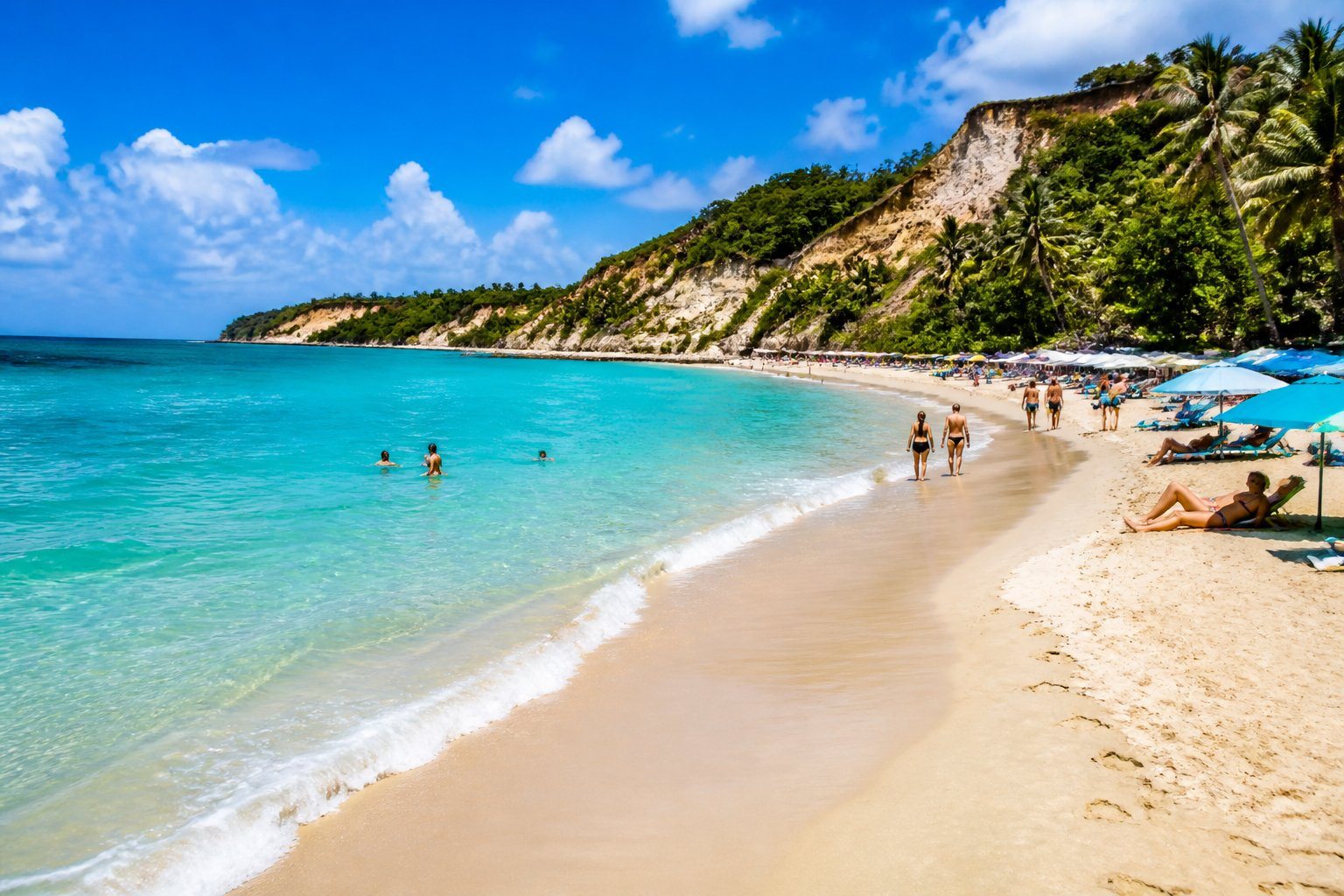 Praia de Pipa com areia dourada, mar azul cristalino, falésias verdes e pessoas aproveitando o dia ensolarado na praia.