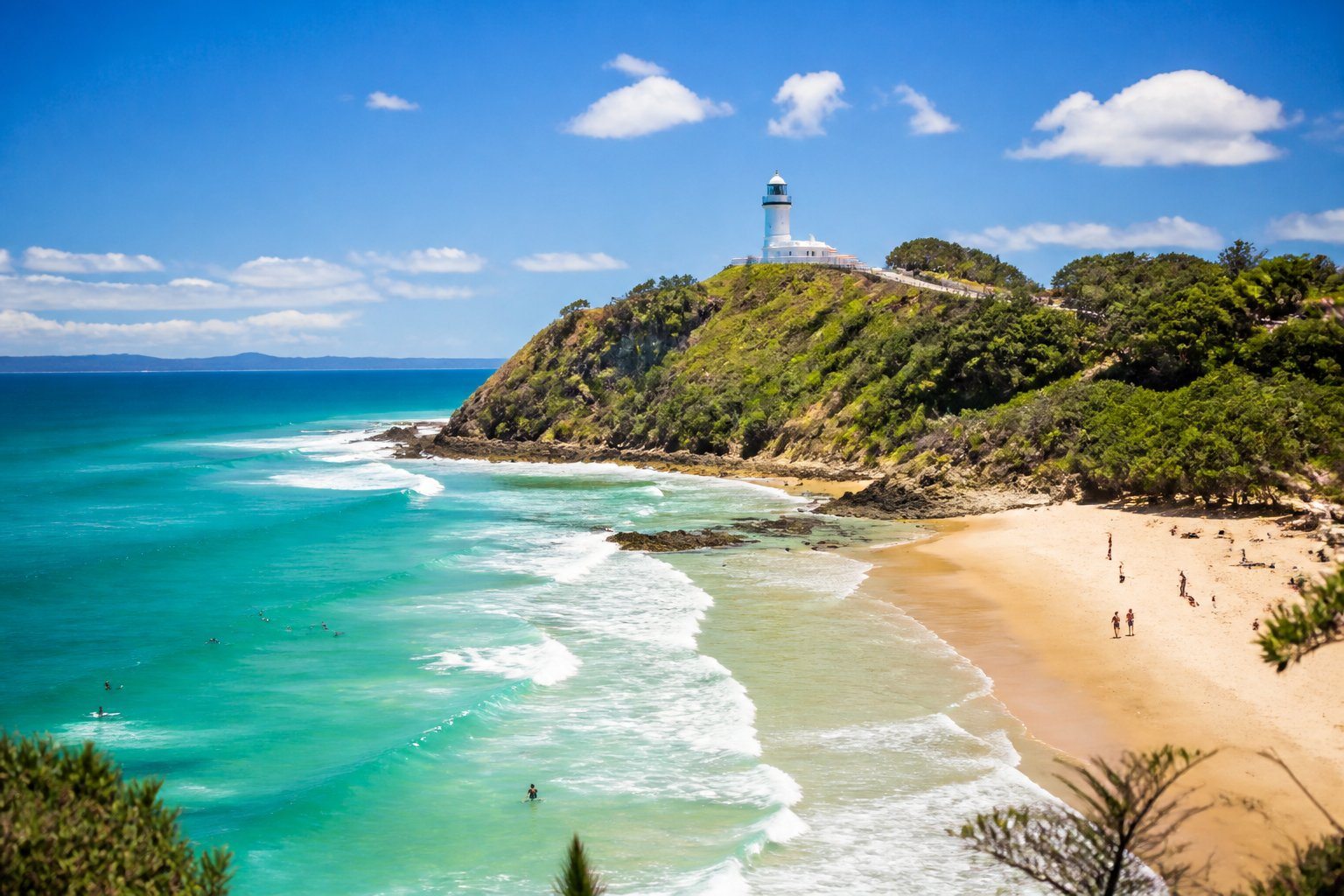 Vista da costa de Byron Bay com praias de areia dourada, águas turquesa, vegetação verde e um farol branco no penhasco ao fundo.