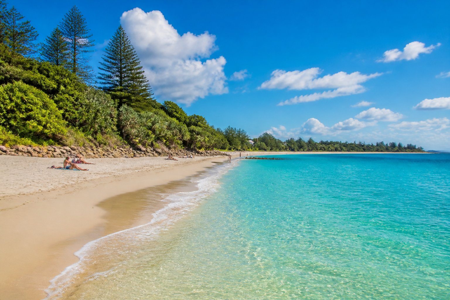 Praia de areia branca com águas cristalinas e vegetação verde ao fundo em Byron Bay.