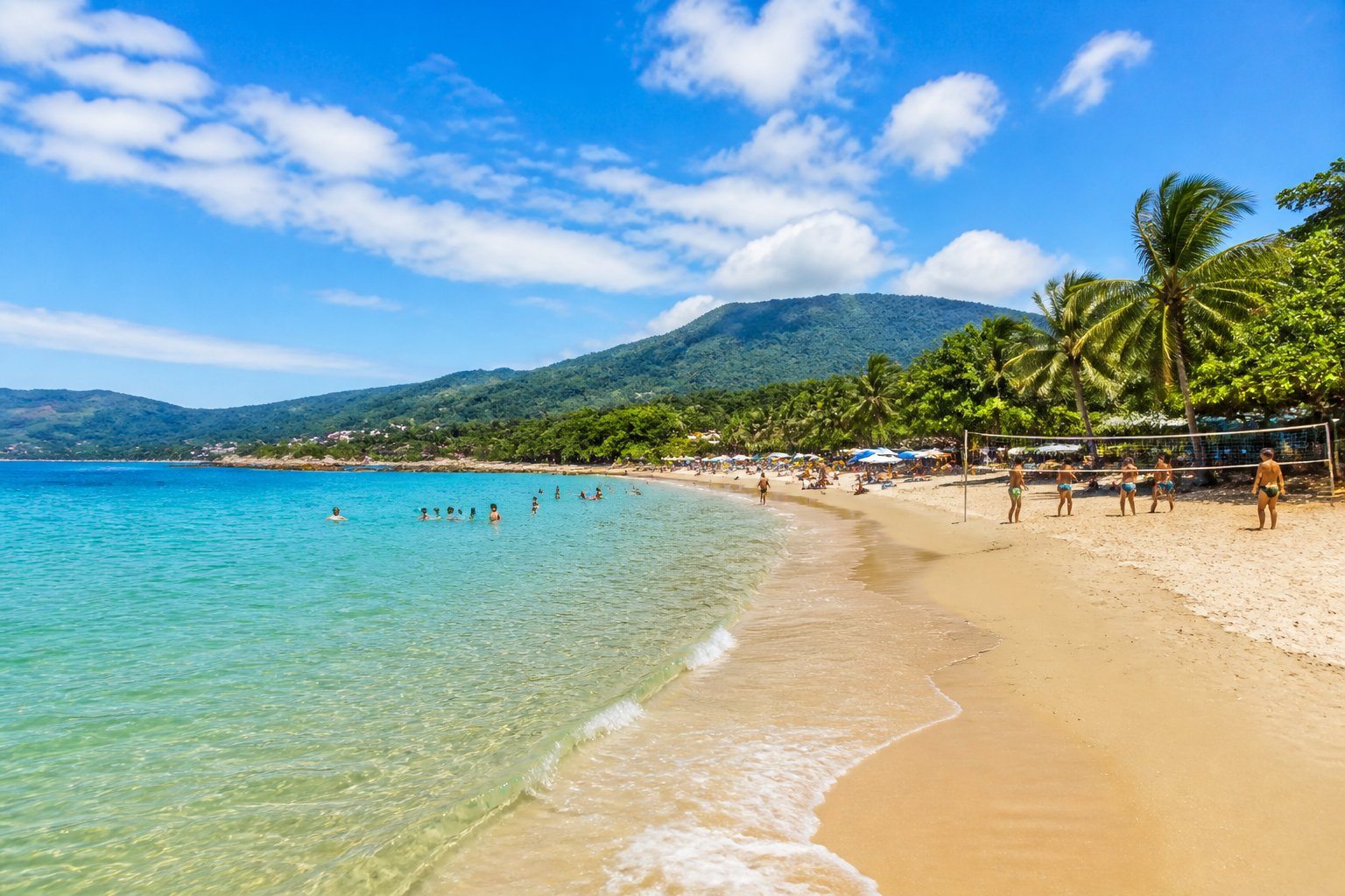 Praia de São Sebastião com areia dourada, mar azul cristalino, vegetação tropical e pessoas aproveitando o dia ensolarado.