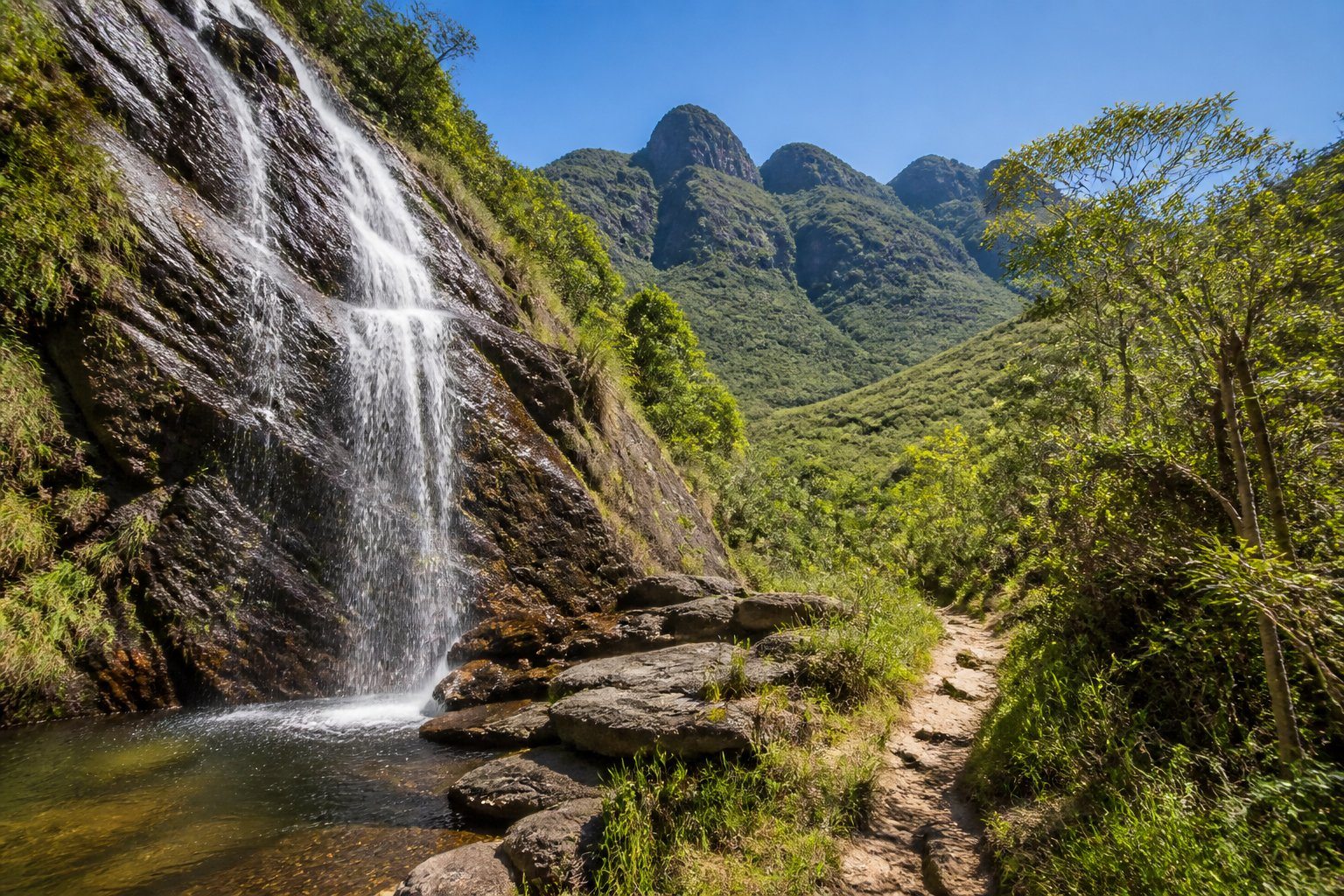 Montanhas verdes com cachoeira caindo em piscina natural e trilha cercada por vegetação densa sob céu azul claro.